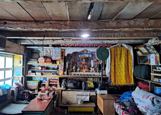 Ein spartanischer, holzgetäfelter Raum mit einem buddhistischen Schrein in der Mitte, umgeben von Regalen mit Büchern und persönlichen Gegenständen, Taschen und Decken