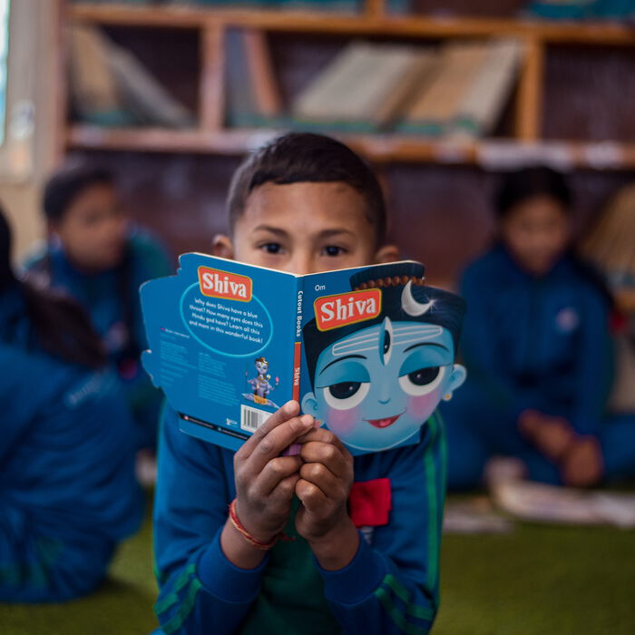 Ein nepalesicher Junge liest in der Schule in einem Buch "Shiva". Im Hintergrund sind weitere Schüler*innen zu sehen.